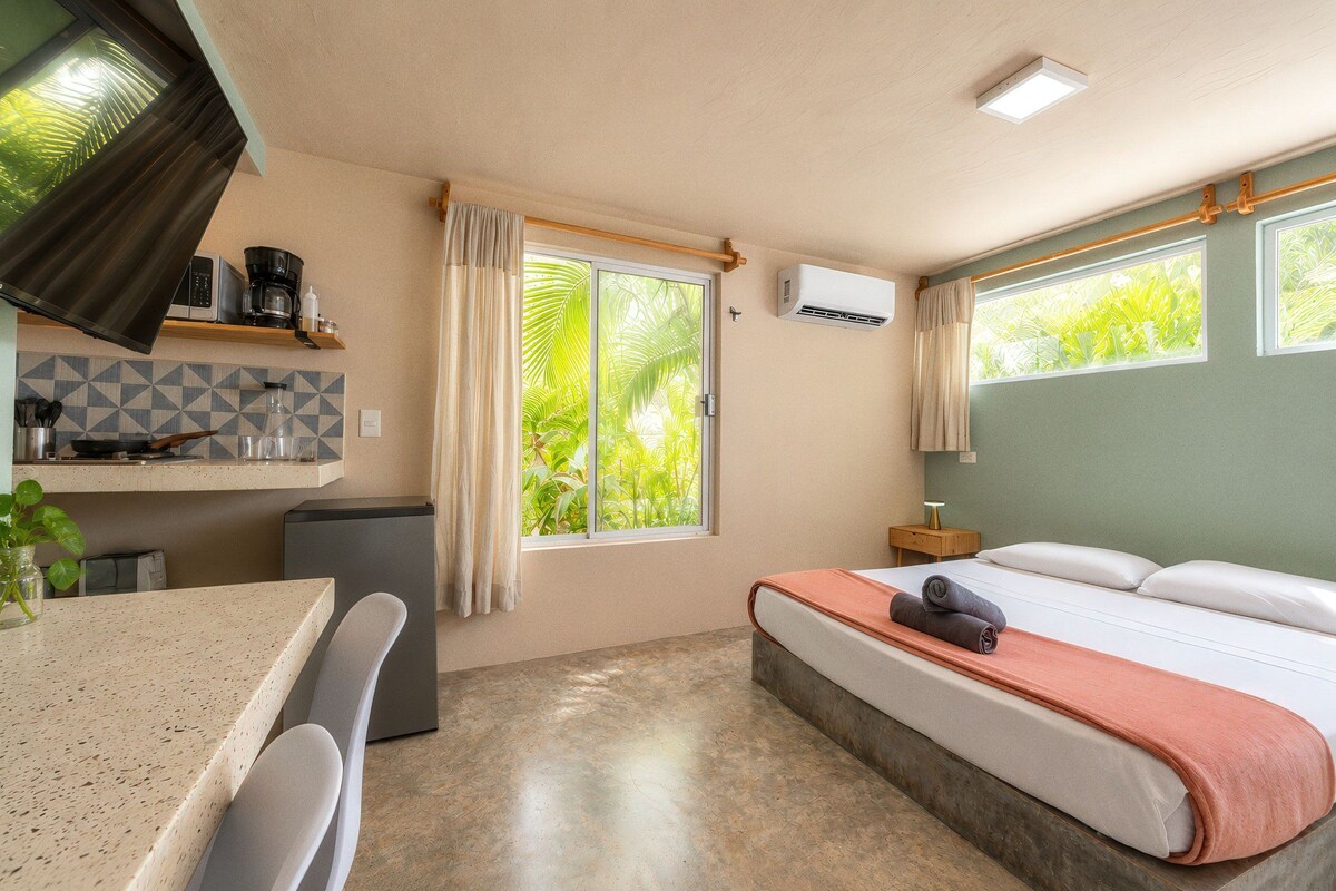Una habitación compacta con cama, kitchenette pequeña, zona de comedor y ventana con vista a follaje.