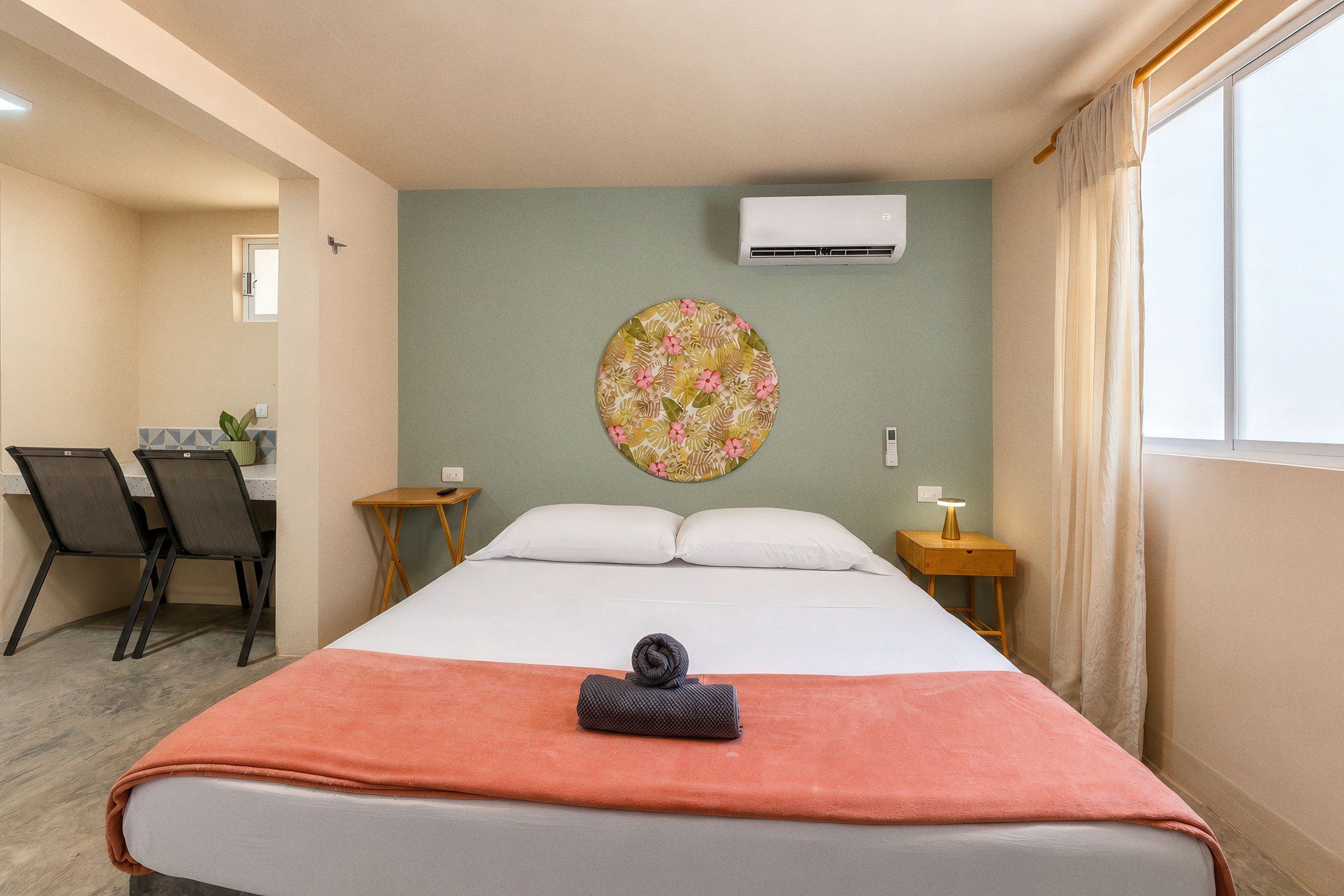 A hotel room with a bed, a desk, two chairs, a small table, an air conditioning unit, and a window with curtains.