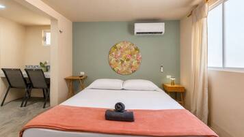A hotel room with a bed, a desk, two chairs, a small table, an air conditioning unit, and a window with curtains.