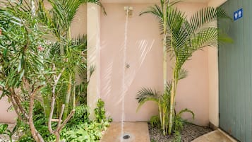 A small outdoor shower area with a water spout, surrounded by tropical plants and a blue sign indicating a men's restroom.