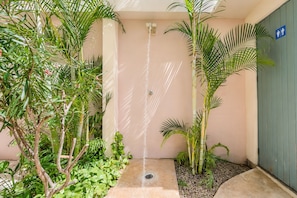 Un área de ducha exterior pequeña con un caño, rodeada de plantas tropicales y un letrero azul que indica un baño de hombres.