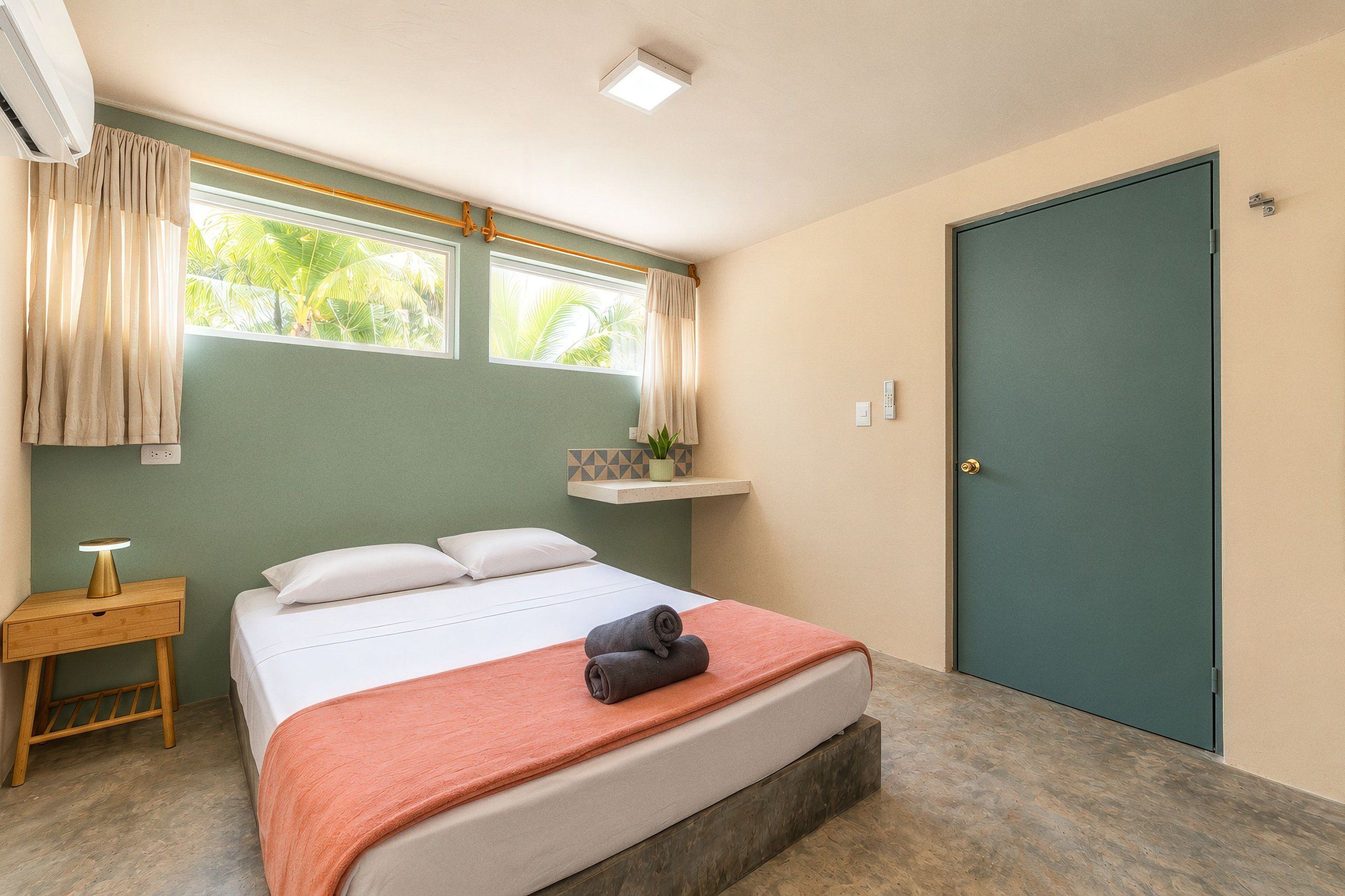 Un dormitorio con cama, mesita de noche, ventana con cortinas, puerta y una pequeña estantería con una planta.