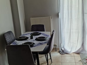 Et spiseområde med et bord dækket til fire personer, stole, en radiator og et vindue med gardiner.