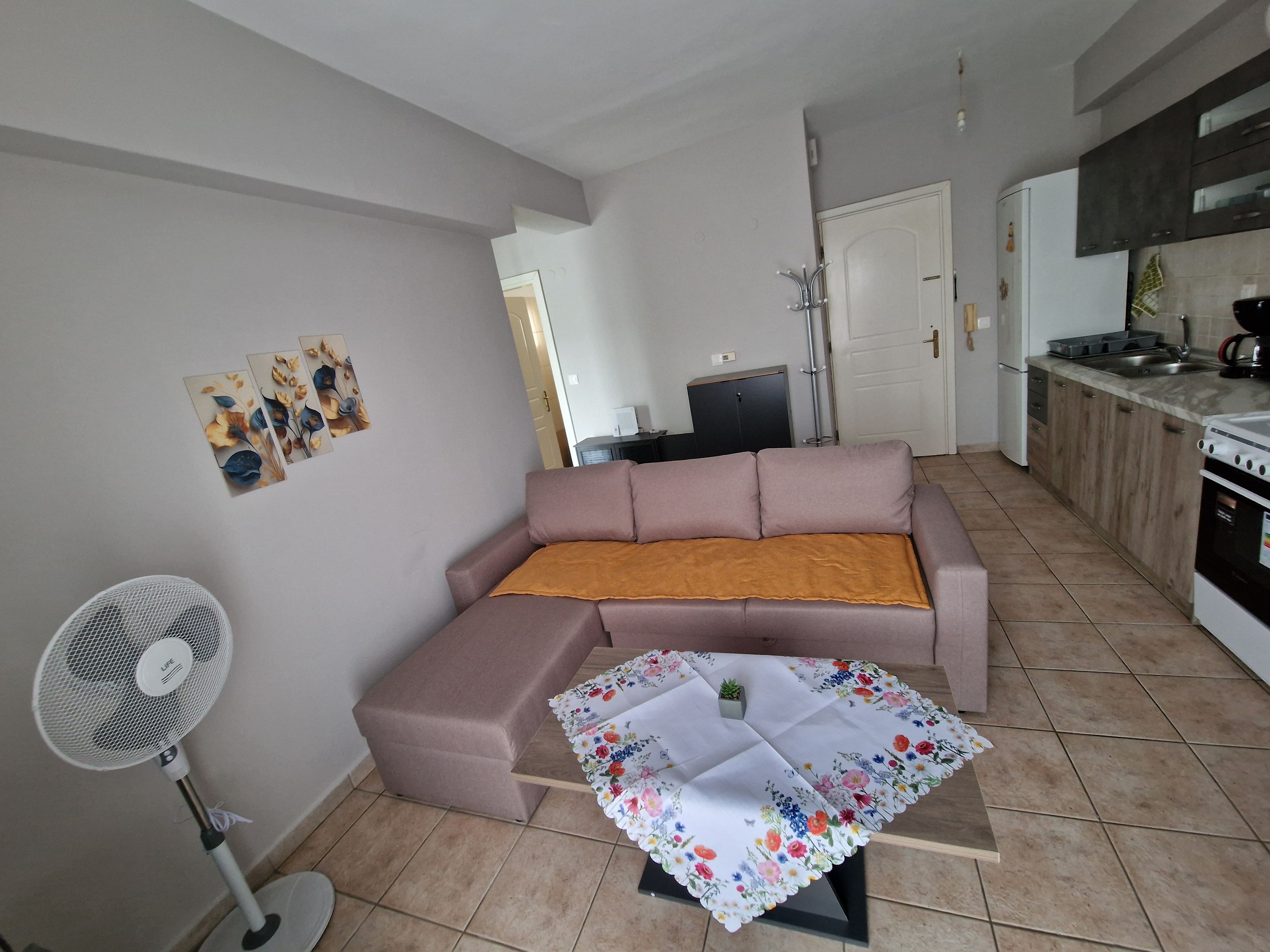 Un salon comprenant un canapé, une petite table recouverte d’une nappe à motifs floraux, un ventilateur sur pied et un coin cuisine avec des placards et des appareils électroménagers.