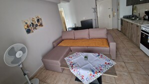 Ein Wohnzimmer mit einer Couch, einem kleinen Tisch mit einer blumigen Tischdecke, einem Standventilator und einer Küchenzeile mit Schränken und Geräten.