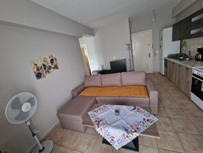 En stue med sofa, et lille bord med blomstret dug, en gulvventilator og et tekøkken med skabe og hårde hvidevarer.