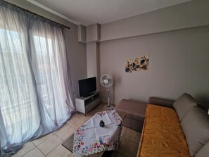 A room with a flat-screen TV on a stand, a standing fan, a floral table, a sofa, and a bed with a brown blanket.