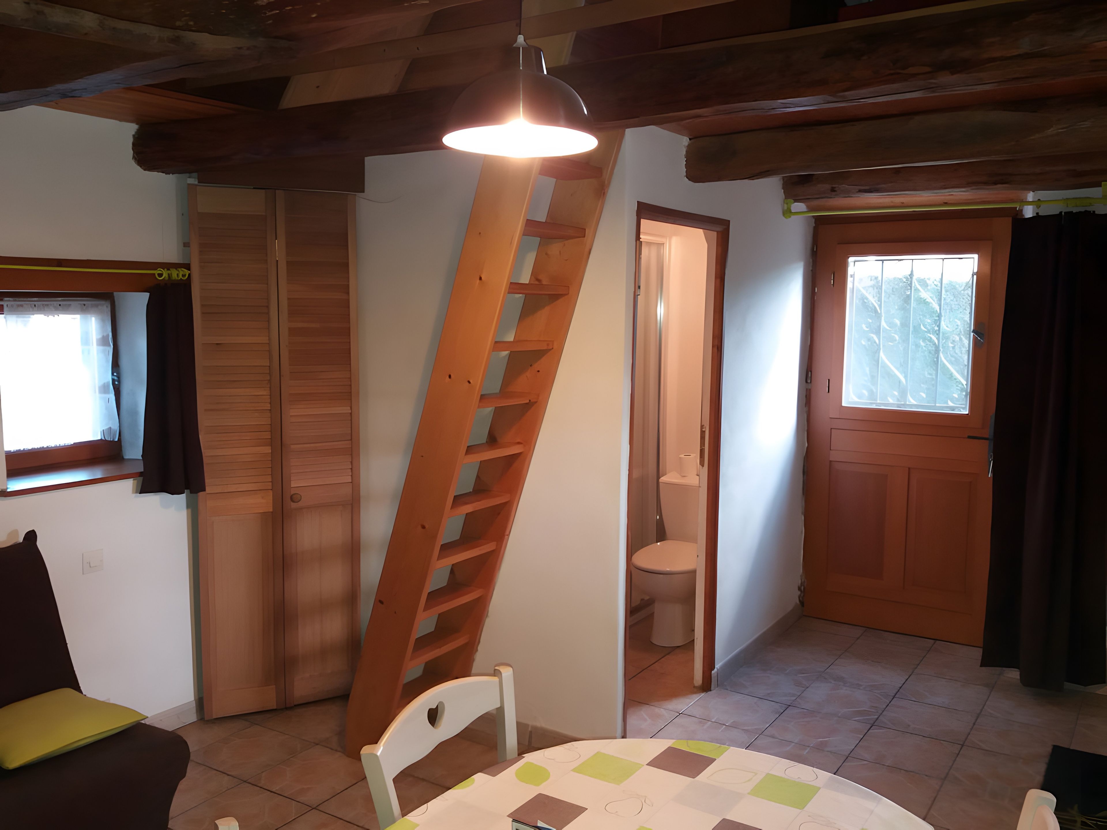 Une pièce avec une échelle en bois, une chaise blanche, une table recouverte d’une nappe à carreaux, une porte en bois et une fenêtre avec des rideaux.