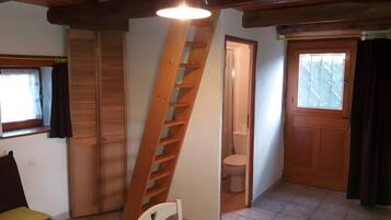 Une pièce avec une échelle en bois, une chaise blanche, une table recouverte d’une nappe à carreaux, une porte en bois et une fenêtre avec des rideaux.