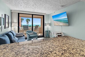 Habitación de hotel con un ventanal amplio que ofrece vista a la playa, una zona de estar con sofá y sillones, y una mesa de comedor.