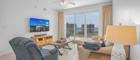 A modern living room with a flat-screen TV, a ceiling fan, a sofa, a coffee table, and a view of the city through large windows.