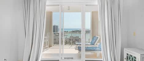 A room with a sliding glass door leading to a balcony with a view of buildings and the sea.