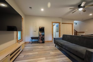 Ein Wohnzimmer mit grauem Sofa, Couchtisch aus Holz, einer Spielkonsole und einem Deckenventilator.
