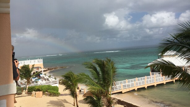 Vista a la playa con valla blanca, palmeras y un edificio con terraza.
