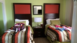 Habitación de hotel con dos camas, cada una con una colcha a rayas, una mesita de noche con lámpara y un espejo en la pared.