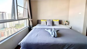 A hotel room with a large bed, a view of the city through a window, and a bedside table with a vase of flowers. - Belle Grand Rama 9 Bangkok (Krung Thep Maha Nakhon)