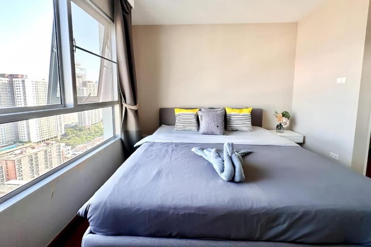 A hotel room with a large bed, a view of the city through a window, and a bedside table with a vase of flowers.