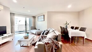 A modern living room with a sofa, a dining table with chairs, a television, and a balcony with curtains. - Belle Grand Rama 9 Bangkok (Krung Thep Maha Nakhon)