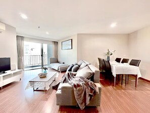 A modern living room with a sofa, a dining table with chairs, a television, and a balcony with curtains. - Belle Grand Rama 9 Bangkok (Krung Thep Maha Nakhon)