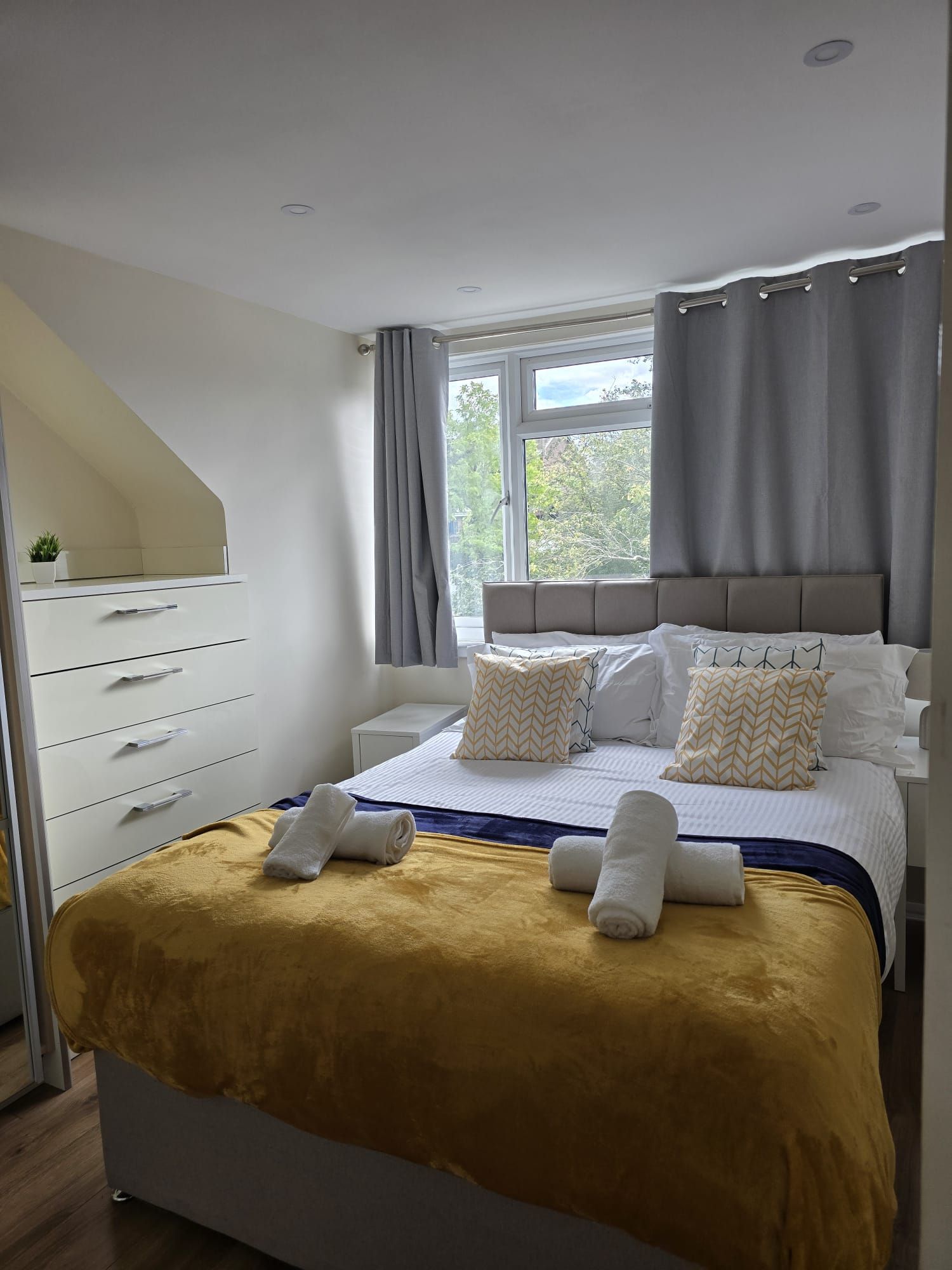 Un dormitorio moderno con una cama, una mesita de noche, un tocador y un armario, junto a una ventana con cortinas.