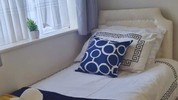 Una cama individual con un cojín blanco y azul estampado, una manta blanca y una toalla enrollada sobre una manta amarilla.