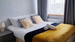 Una cama con edredón blanco, dos almohadas, un plaid dorado y una mesita de noche con lámpara.