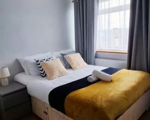A bed with a white duvet, two pillows, a gold throw, and a bedside table with a lamp. - Stylish 4BR Home in Essex | Sleeps 7 (South Benfleet)