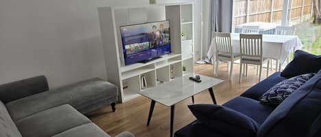 Una sala de estar con televisor, sofás grises, una mesa de centro y un comedor con mesa y sillas.