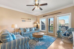 A living room with striped sofas, a round coffee table, a ceiling fan, and a view through the glass doors. - Pointe of No Return: Semi Oceanfront Condo (Kill Devil Hills)