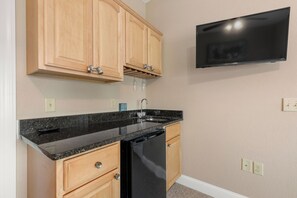A compact kitchen with wooden cabinets, a black countertop, a dishwasher, and a mounted flat-screen TV. - Pointe of No Return: Semi Oceanfront Condo (Kill Devil Hills)