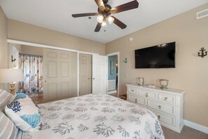 A bedroom with a bed, a dresser, a ceiling fan, a television, and a door leading to another room. - Pointe of No Return: Semi Oceanfront Condo (Kill Devil Hills)