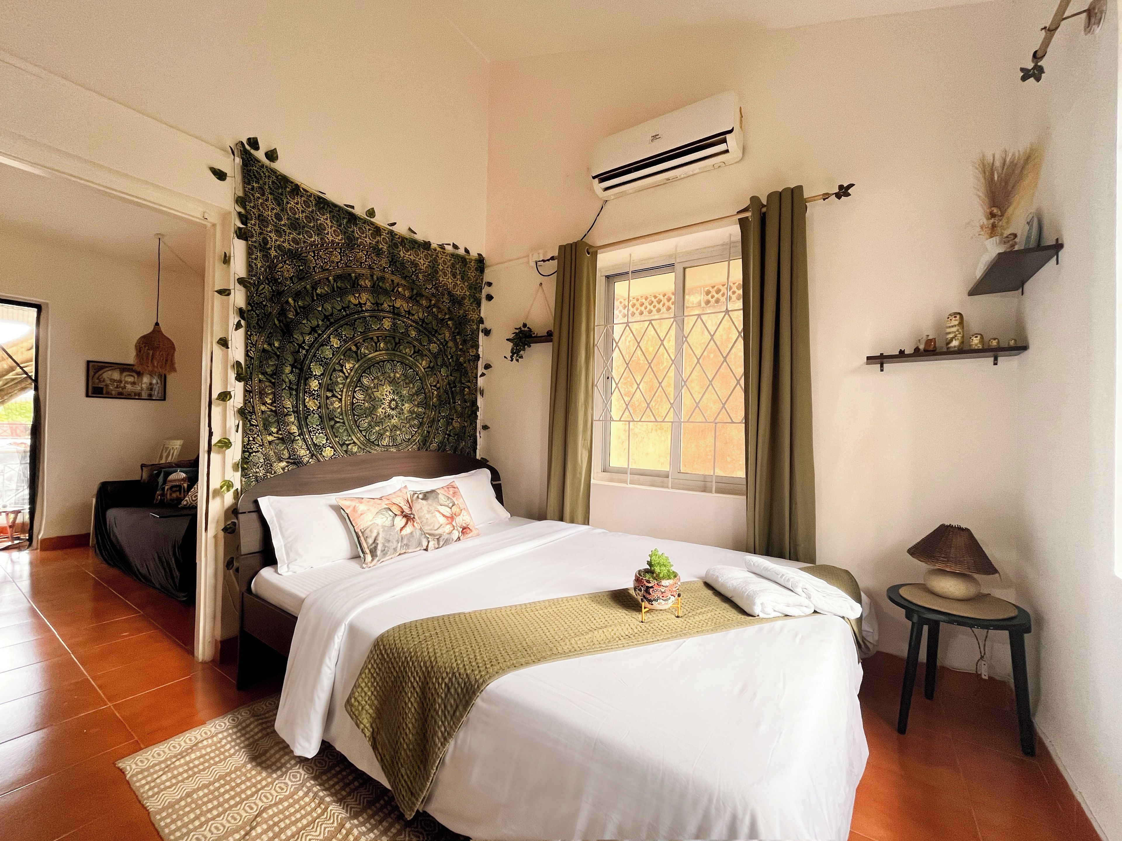 A bedroom with a bed, a window with curtains, an air conditioning unit, a small table with a lamp, and a decorative wall tapestry.