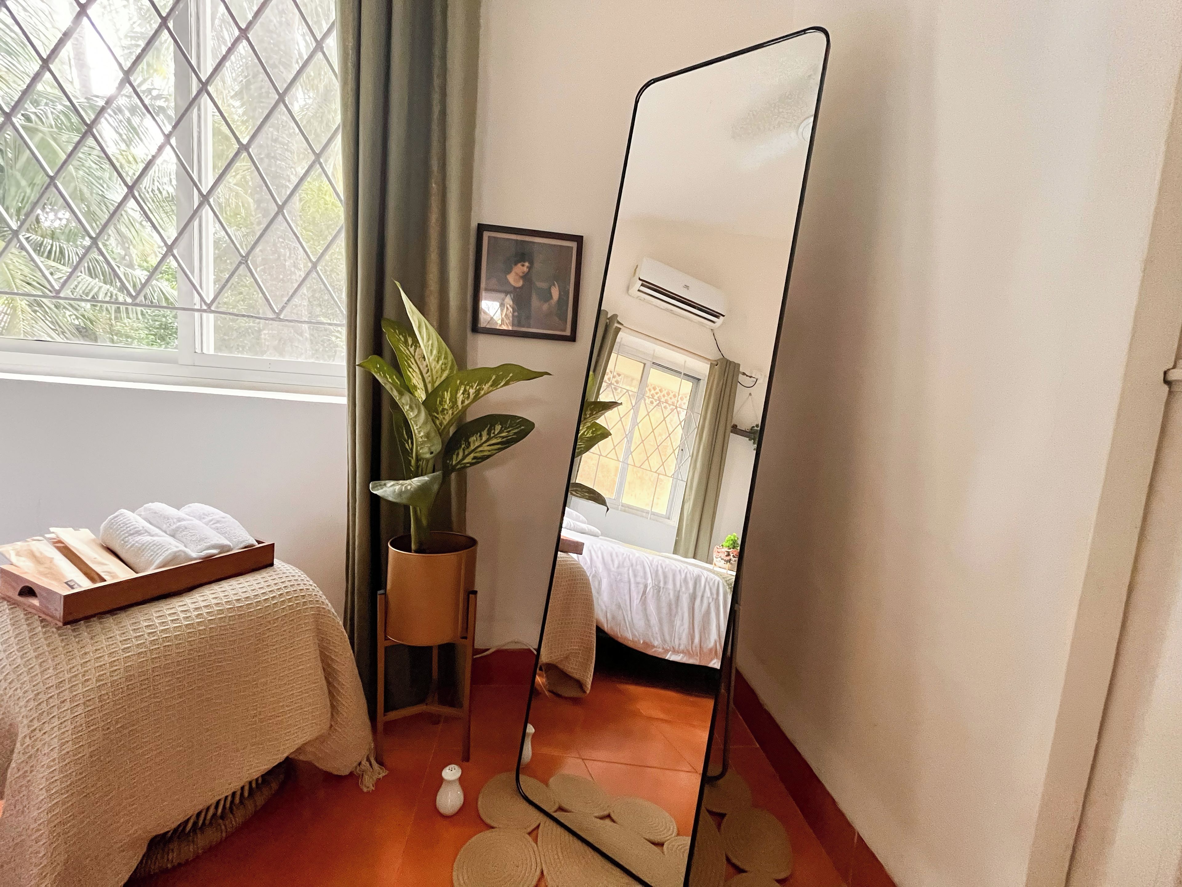 A room with a bed, a potted plant, a mirror, and a window with curtains.
