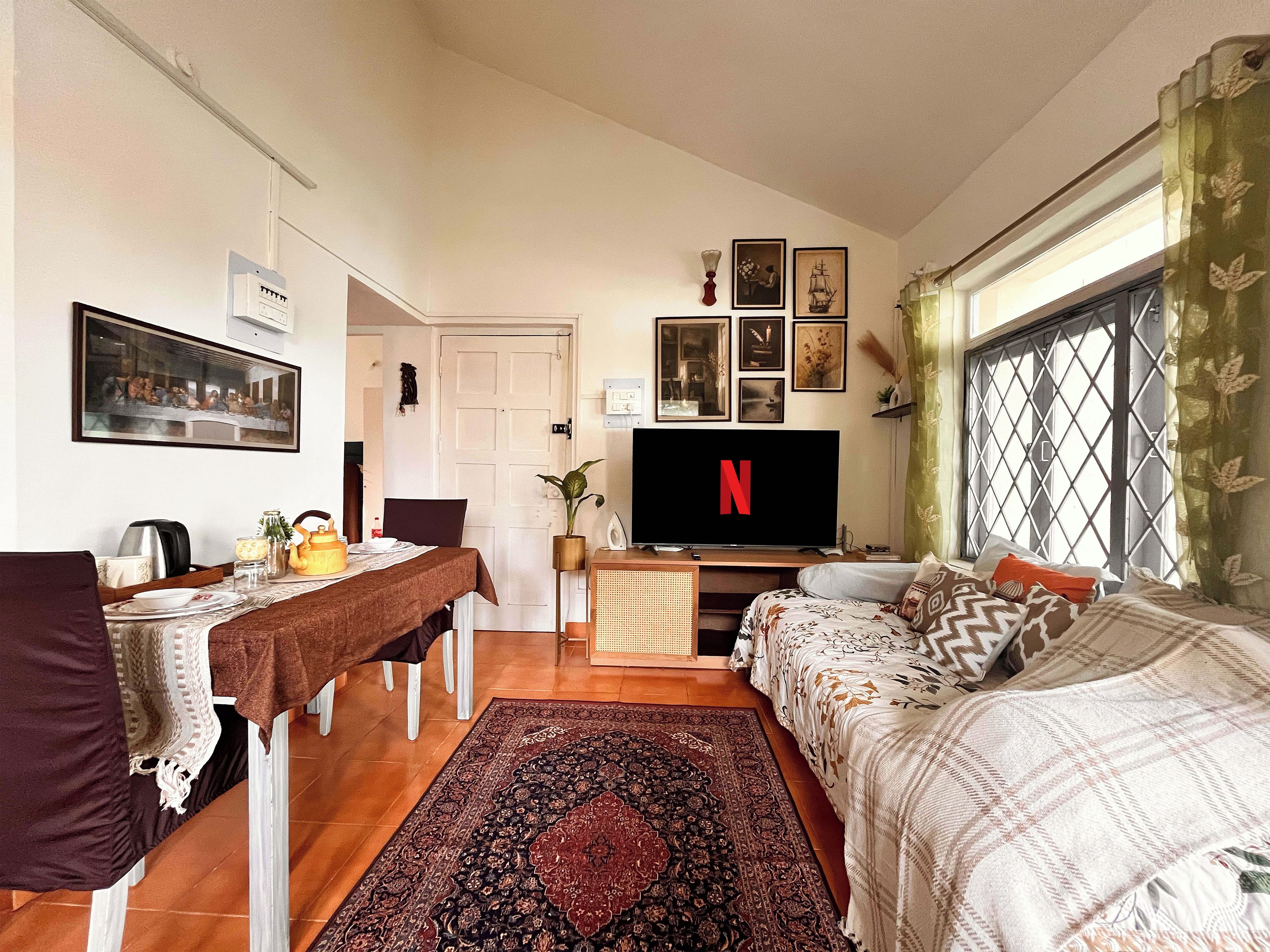 A cozy living room with a television, a dining area with a table and chairs, and a large window with curtains.