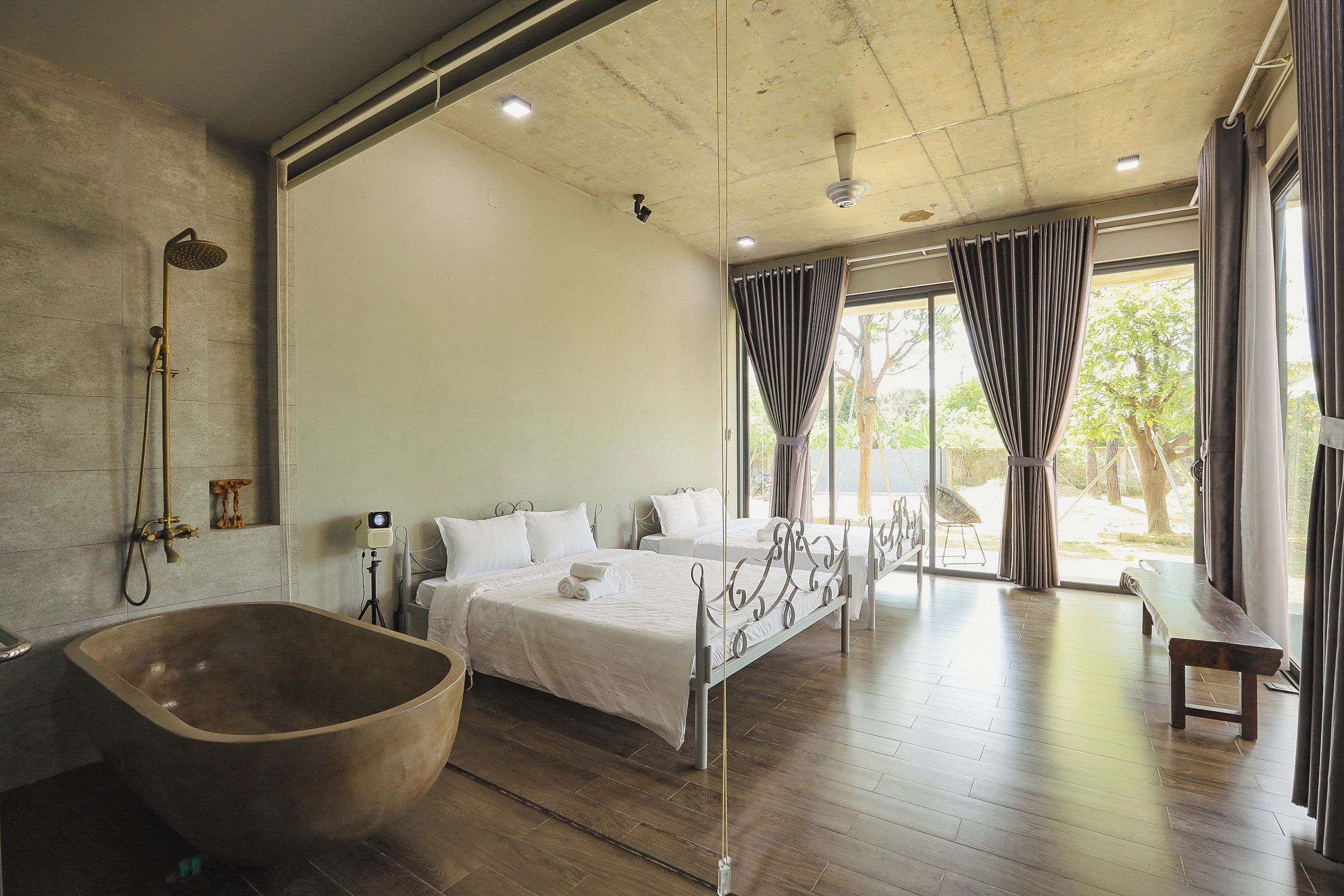 A modern bedroom with a large wooden bathtub, a bed with white linens, and a view of the outdoors through large windows.