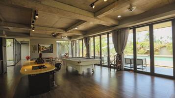 A spacious room with a pool table, a bar area, and large windows offering a view of the outdoors.
