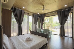 Habitación de hotel con cama, ventilador de techo y ventanas amplias con cortinas.