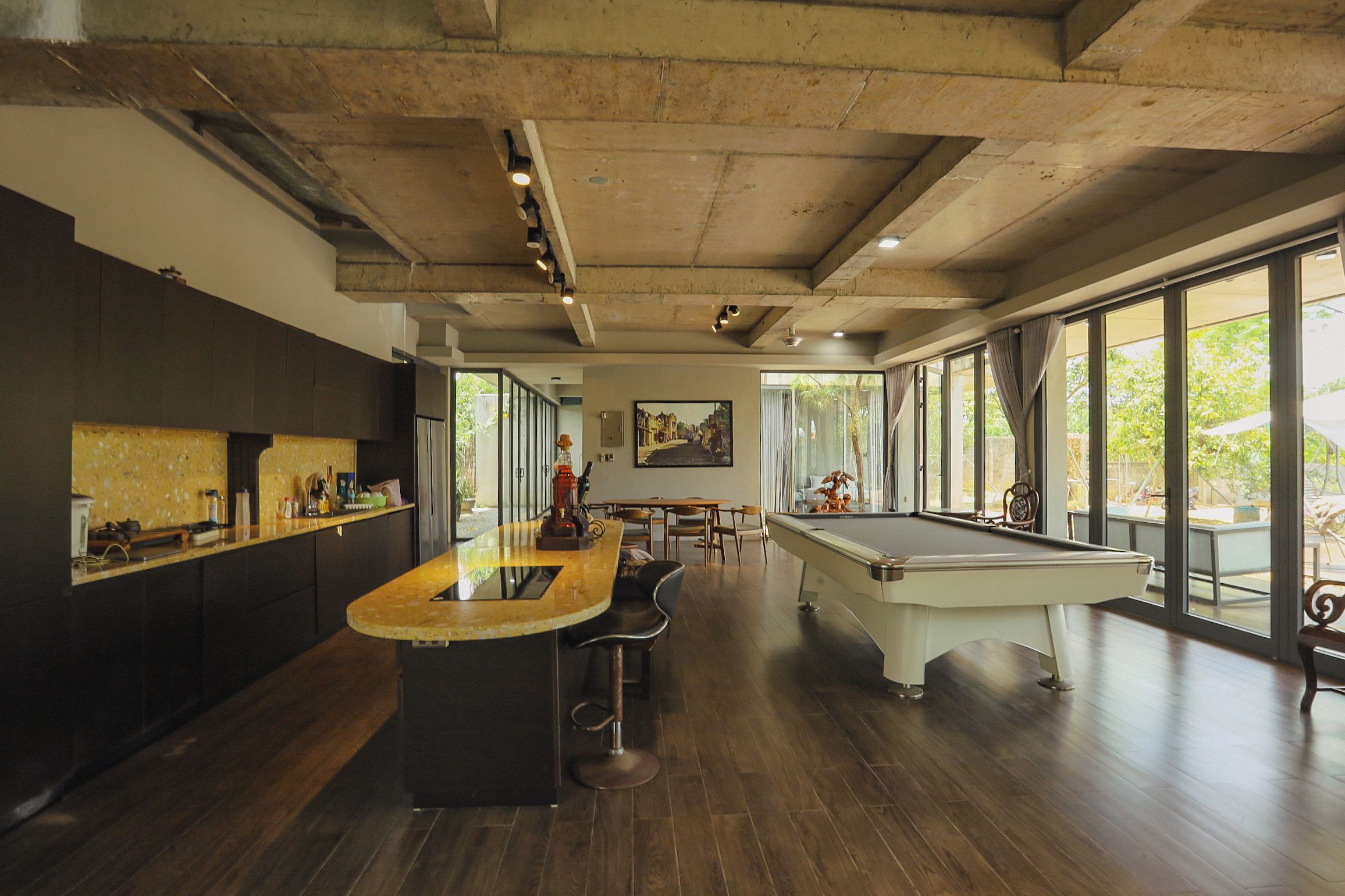 A spacious room with a pool table, a bar area with stools, and large windows offering a view of the outdoors.