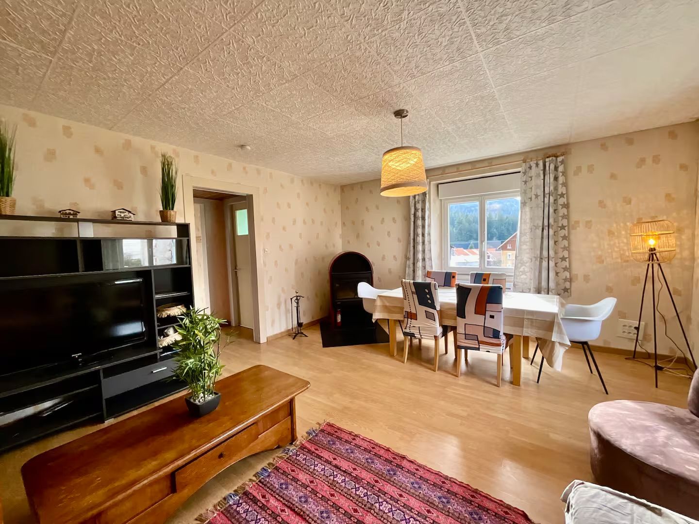 A living room with a flat-screen TV, a wooden coffee table, a patterned rug, a dining area with a table and chairs, and a window with a view.