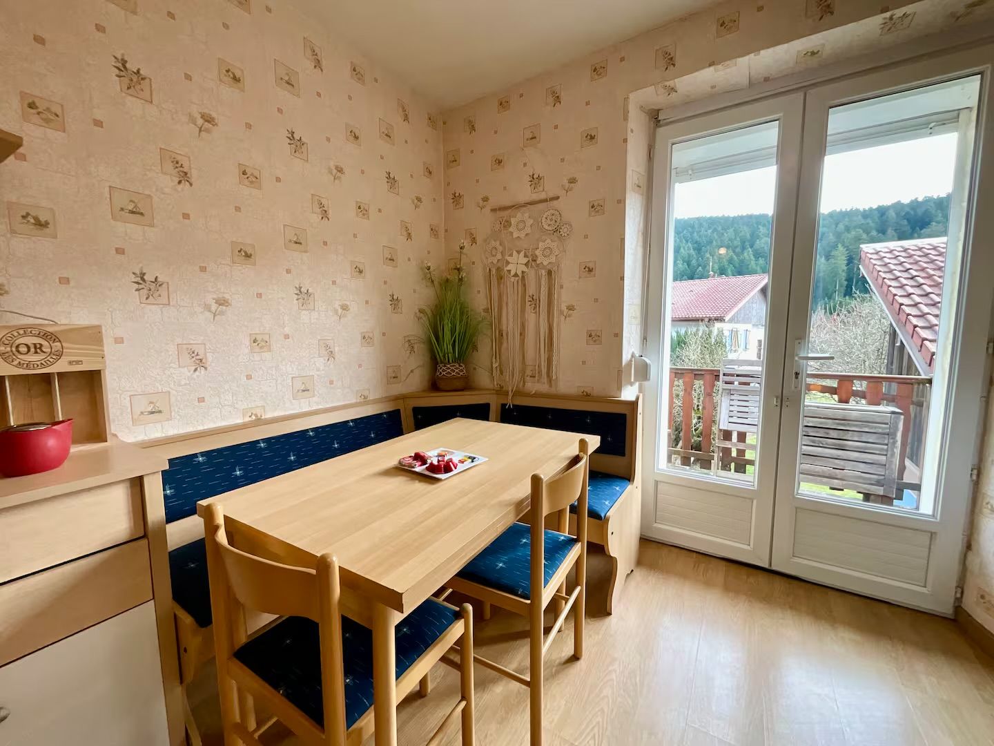 A room with a wooden table and chairs, a bench, a red bowl, a plant, and a view through a glass door.