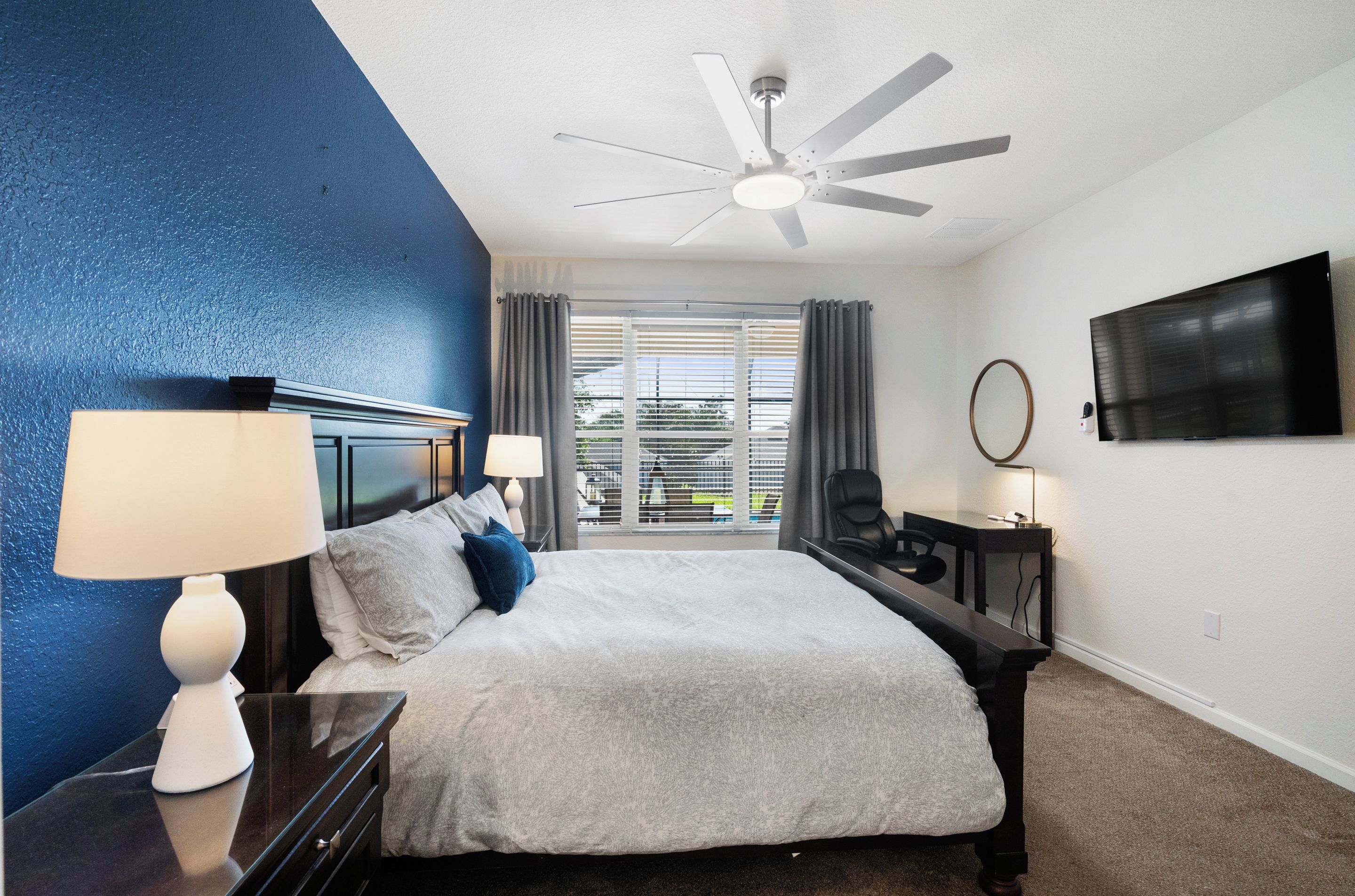 A hotel room with a bed, a desk, a chair, a TV, a ceiling fan, and a view of the outdoors through the window.