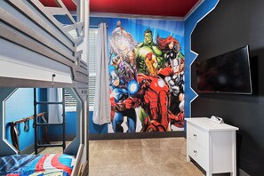 Ein Schlafzimmer mit Etagenbett, einer großen Wandmalerei von Superhelden, einem Fernseher und einer weißen Kommode.