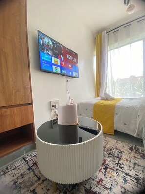 A hotel room with a flat-screen TV mounted on the wall displaying various streaming services, a round table with a remote control, and a bed with a yellow bedspread. - Studio apartment at Tsavo sunset (Kiambu)