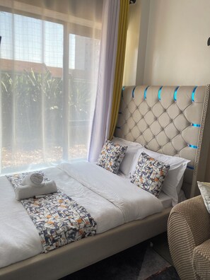 A hotel room with a bed, a window with curtains, and a view of greenery outside. - Studio apartment at Tsavo sunset (Kiambu)