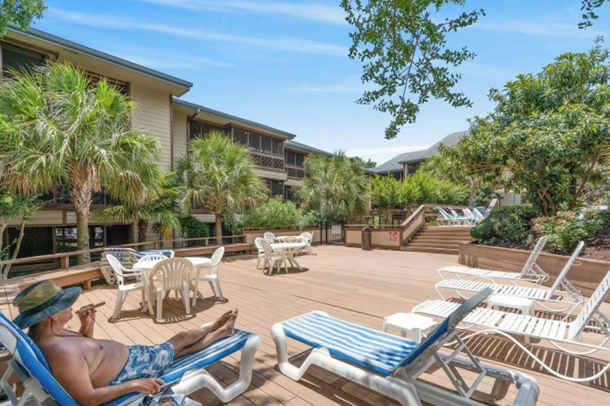 Un espace aménagé au bord de la piscine avec des chaises longues, un homme qui se détend et un bâtiment doté de balcons.