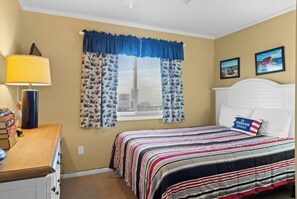A bedroom with a bed, a nightstand, a lamp, a window with curtains, and two wall-mounted pictures. - Pelican's Landing 208- 3BR in Shore Drive- Pool (Myrtle Beach)