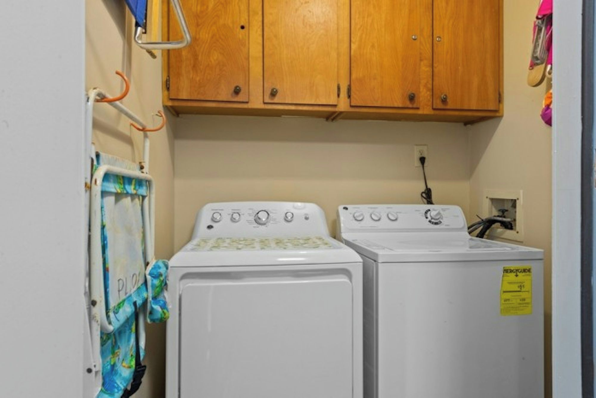 A laundry room with two white washing machines, wooden cabinets above, and a yellow sign on the right machine.