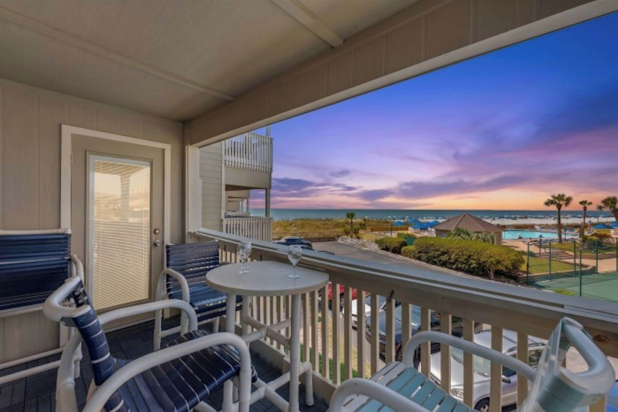 A balcony with a view of the ocean and a sunset. There are chairs and a table with a glass.