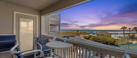 A balcony with a view of the ocean and a sunset. There are chairs and a table with a glass.
