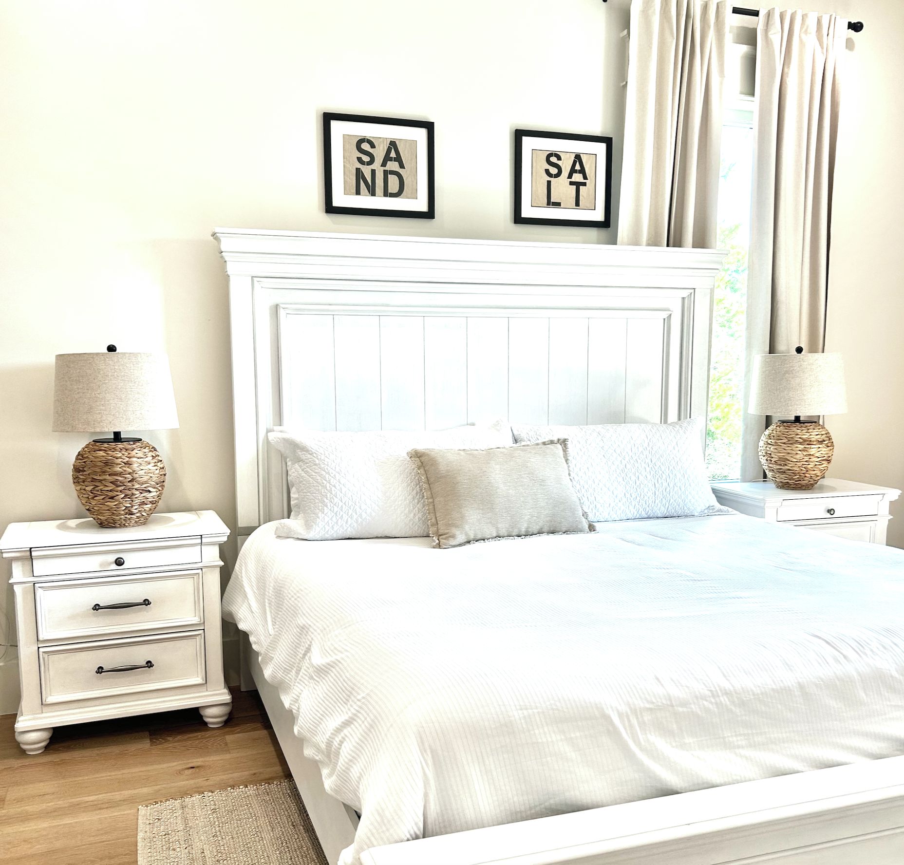 A bedroom with a white bed, two nightstands with lamps, a white dresser, and two framed signs on the wall.